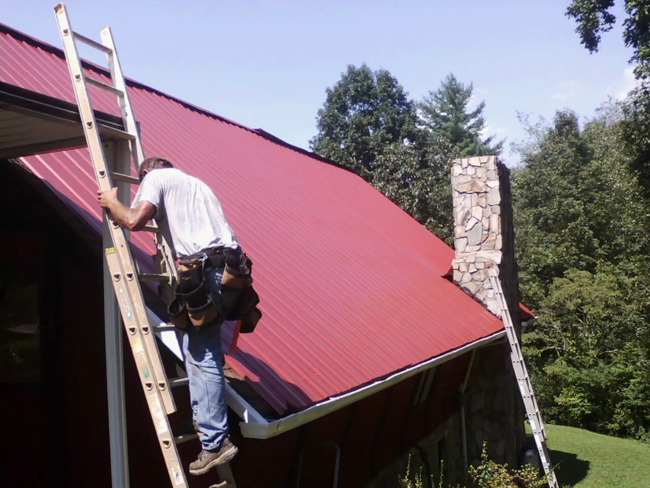 Roofer on a ladder beside a red metal roof and stone chimney — Soffit & Fascia Repair by Roofing Brothers in Morris Plains